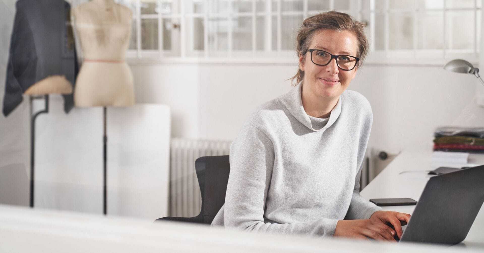 Kvinde med briller og lys sweater sidder ved sin bærbare computer i et lyst atelier med buster og stof i baggrunden.