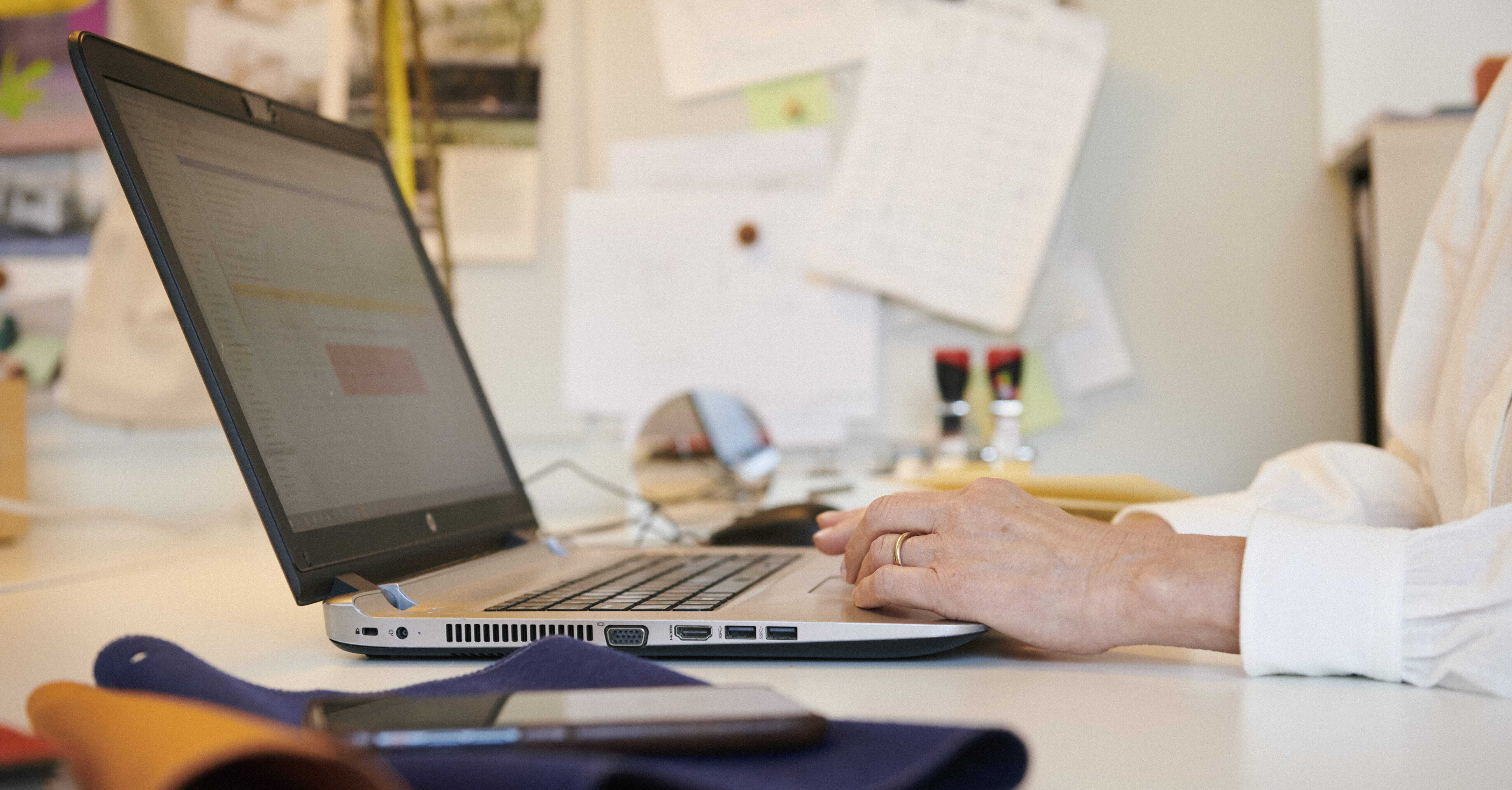Medarbejder arbejder ved skrivebord med bærbar computer, notater og stempler i baggrunden.