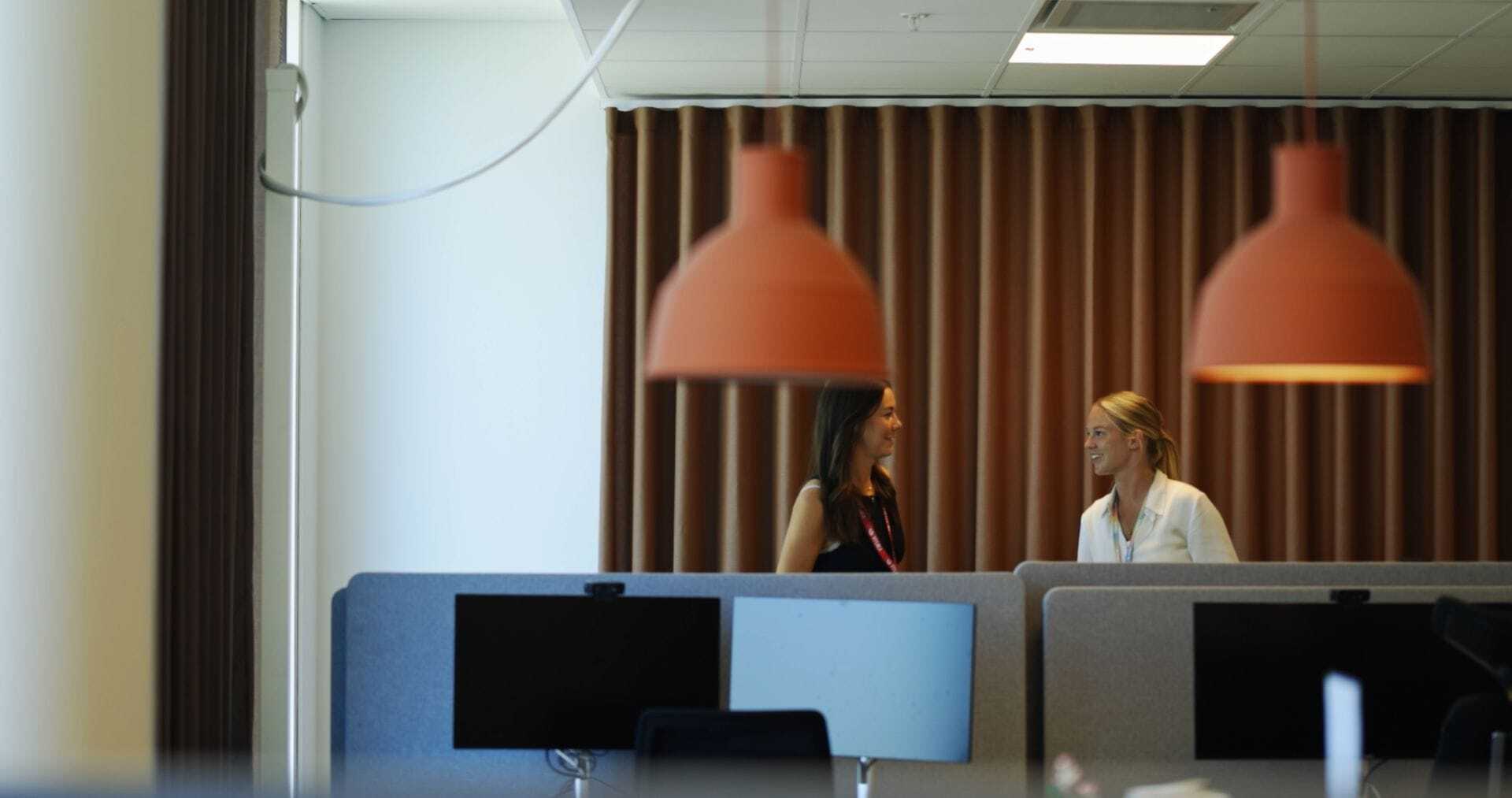 To kolleger i samtale ved skrivebordspladser på et kontor, med orange pendellamper og gardiner i baggrunden.