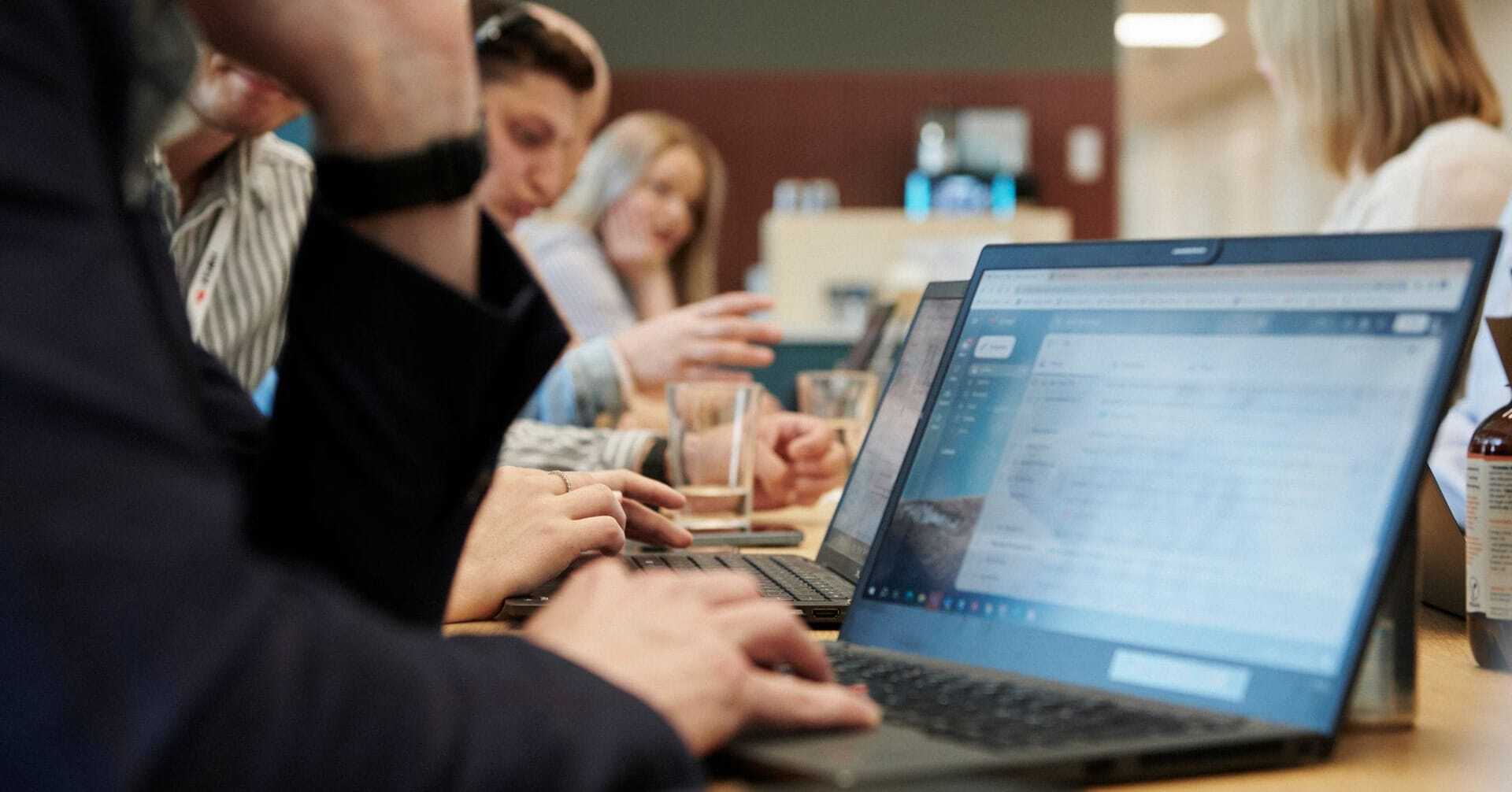 Person arbejder på en bærbar computer ved et bord med flere kolleger i baggrunden.